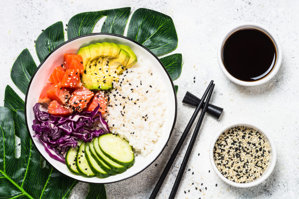 Salmon poke bowl with rice and vegetables on white stone table with tropical leaves.